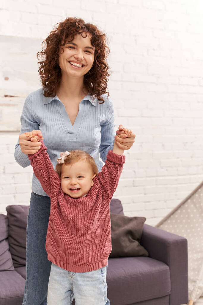 loving motherhood, balancing work and life, family bonding, happy and curly mother lifting toddler daughter, togetherness, cozy living room, quality time, denim jeans, engaging with child  - Photo, Image