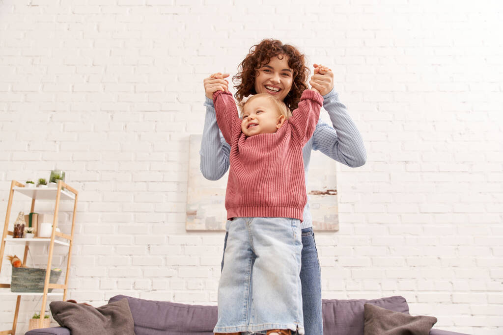 engaging with child, balancing work and life, family bonding, happy and curly mother lifting toddler daughter, togetherness, cozy living room, quality time, denim jeans, loving motherhood  - Photo, Image