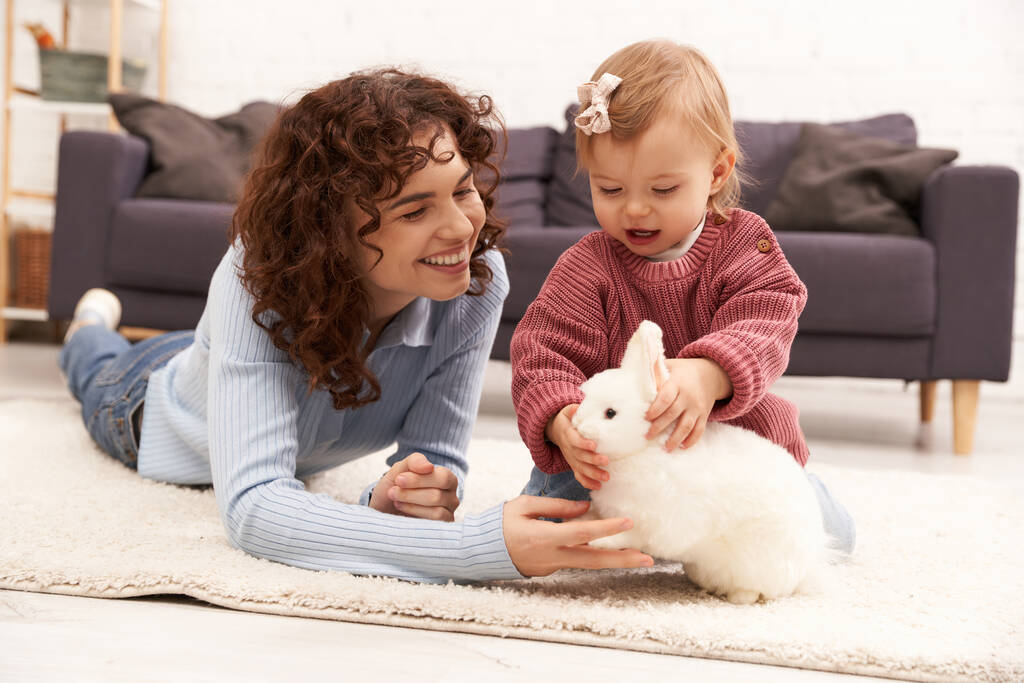 angażowanie się z dzieckiem, wesołą i kręconą kobietą leżącą na dywanie z córeczką malucha w przytulnym salonie, zabawa z królikiem, jakość czasu rodzinnego, luźny strój, wiązanie matki z dzieckiem  - Zdjęcie, obraz