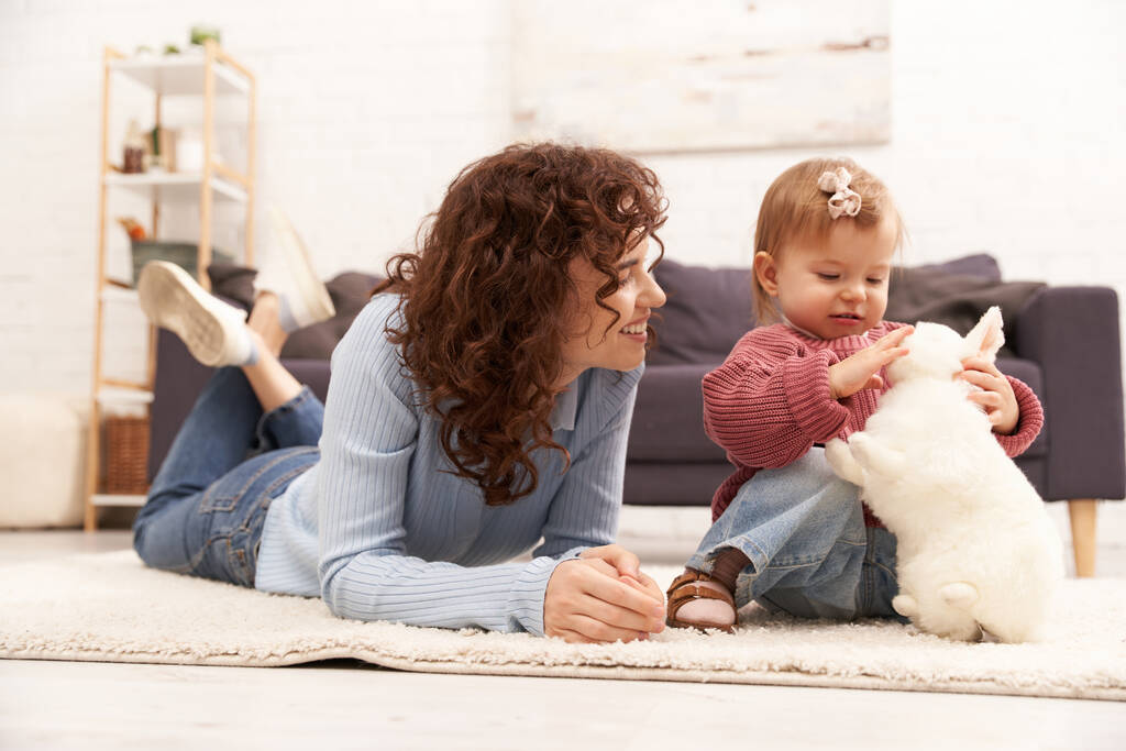 Einbeziehung mit Kind, glückliche Frau auf Teppich liegend mit Kleinkind-Tochter im gemütlichen Wohnzimmer, Spielen mit Kaninchen, hochwertige Familienzeit, lässige Kleidung, Bindung zwischen Mutter und Kind, Mutterschaft  - Foto, Bild