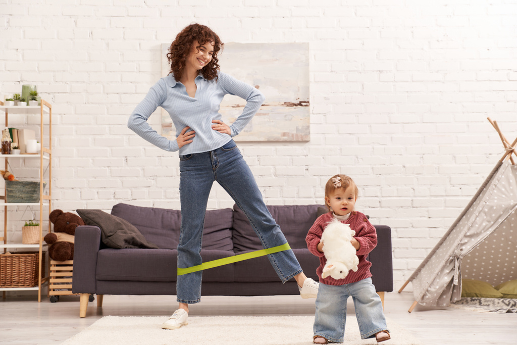 busy mom, home workout, sport, working mother, balanced lifestyle, happy woman exercising with resistance band near toddler daughter with soft toy in cozy living room, modern parenting  - Photo, Image