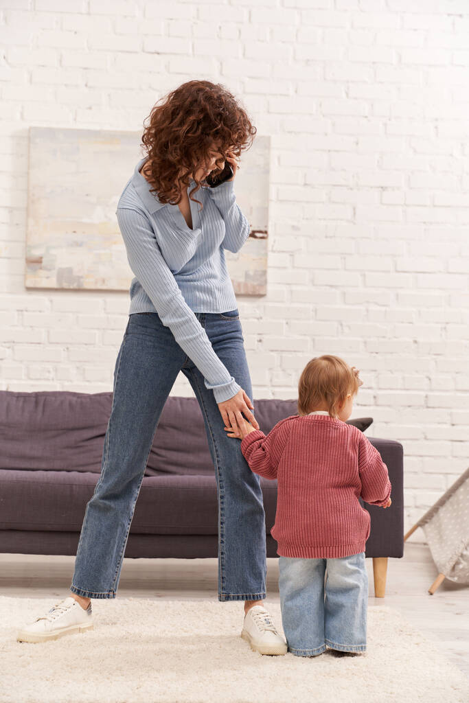 full length of toddler baby doing her first steps near curly mother, engaging with kid, denim jeans, casual attire, family time, modern parenting, work life balance, cozy living room  - Photo, Image