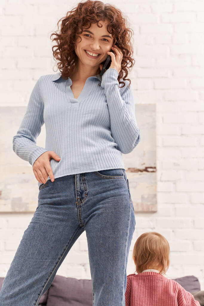 cheerful young woman talking on smartphone standing near toddler kid, balancing between work and life, modern parenting, remote work, career and family, casual attire  - Photo, Image