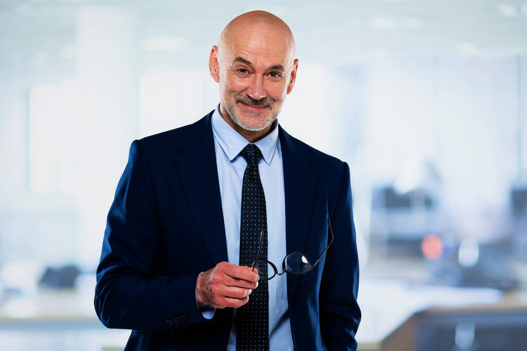 Executive businessman wearing suit and tie while standing at the office. Professional male looking at camera and smiling. He is holding glasses in his hand.  - Photo, Image