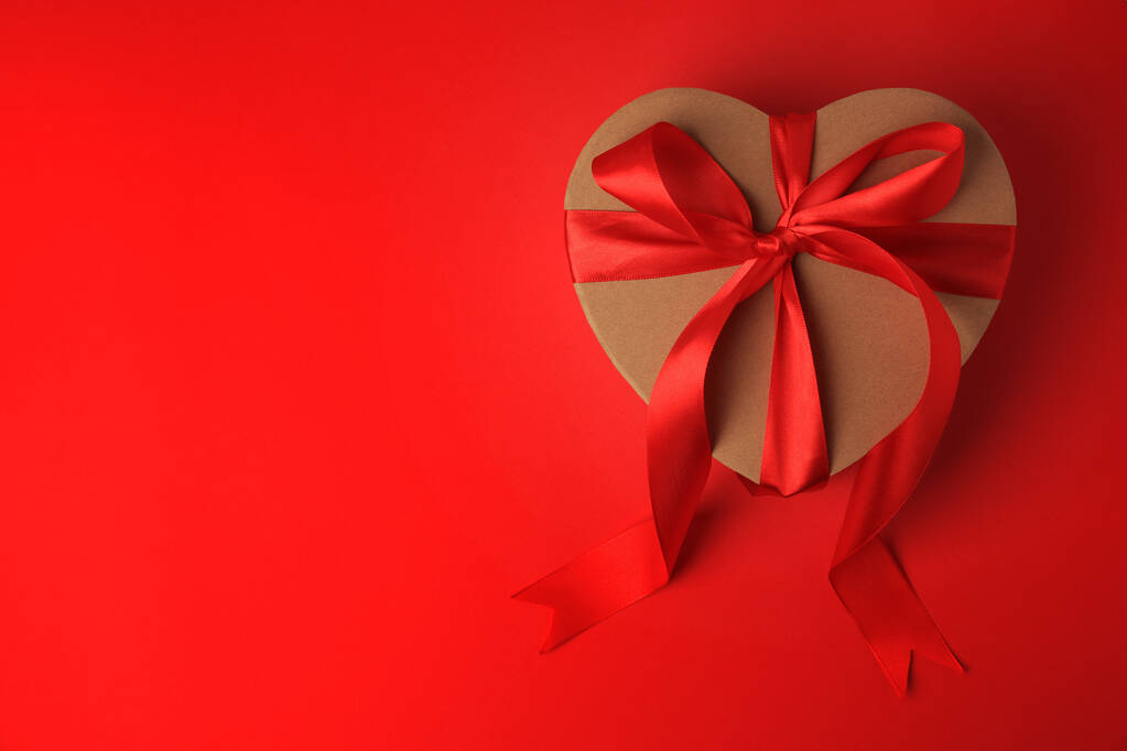Подарочная коробка в форме сердца с бантом на красном фоне, вид сверху. Пространство для текста - Фото, изображение
