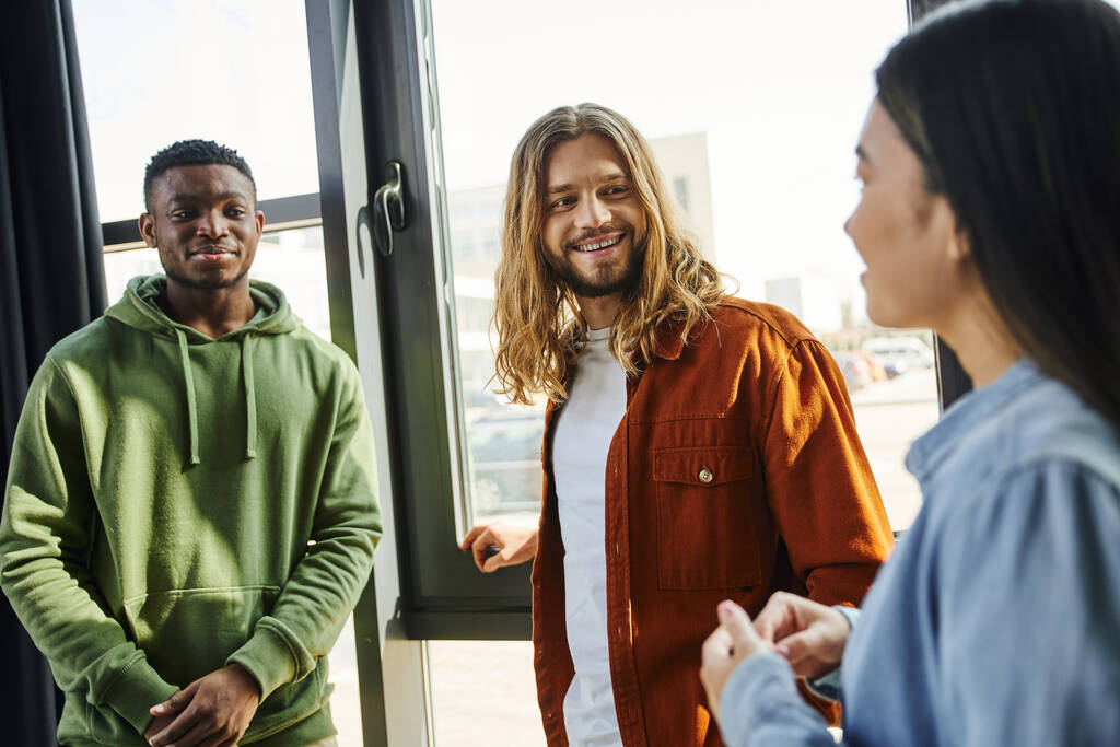 creative multicultural business people talking and planning startup project in modern office space, long haired and stylish man smiling near african american colleague and asian woman - Photo, Image