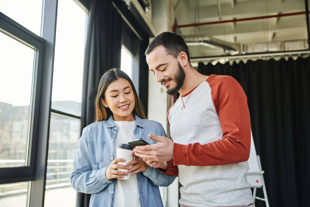 счастливый бородатый мужчина показывает мобильный телефон привлекательной азиатской женщине, держащей кофе, чтобы пойти в офис, коллеги по межрасовому бизнесу, молодые предприниматели, сотрудничество стартап-проекта - Фото, изображение