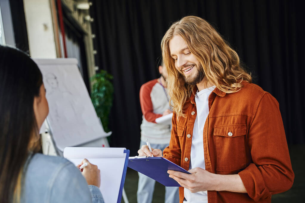 stylish and long haired man smiling while writing on clipboard near woman and medical instructor with flip chart on blurred background, first aid seminar, health care and emergency response concept - Photo, Image