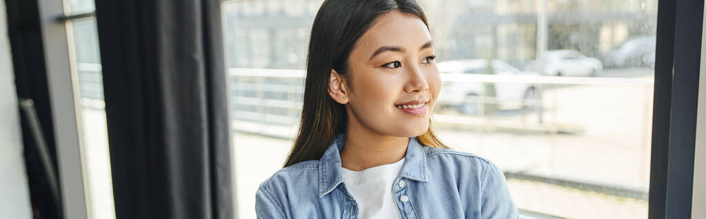 atraktivní a radostná asijská žena v modré džínové košili, s brunetkou vlasy stojí u okna v moderní kanceláři a odvrací pohled, mladý a úspěšný podnikatel, banner - Fotografie, Obrázek