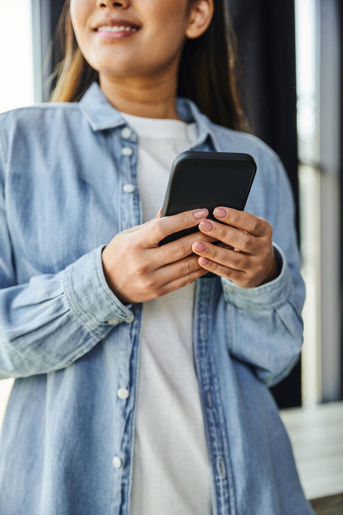 обрезанный вид улыбающейся бизнесвумен в синей джинсовой рубашке в сети на мобильном телефоне, стоя в современном офисе на размытом фоне, молодой предприниматель, успешная молодежь - Фото, изображение