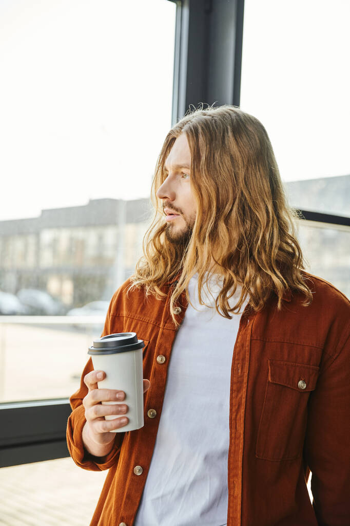 entrepreneur rêveur, aux cheveux longs et barbu dans des vêtements décontractés tenant tasse en papier avec café, debout près de la fenêtre et regardant loin, cool homme de style hipster dans le bureau moderne - Photo, image