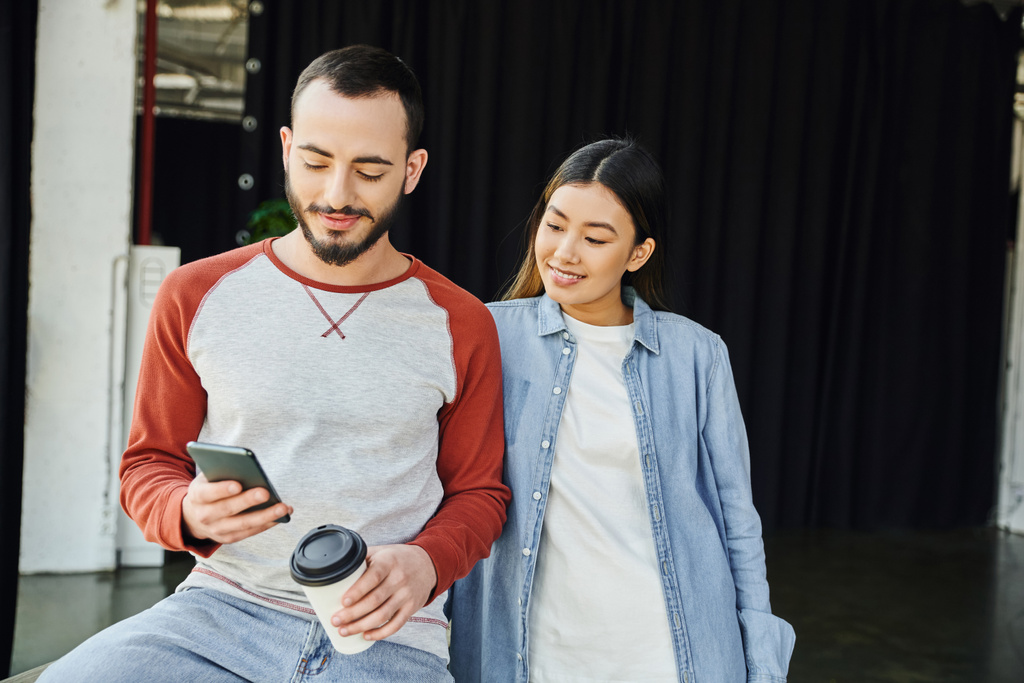 позитивный бородатый мужчина держит бумажную чашку с кофе, чтобы пойти и просматривать Интернет на смартфоне рядом улыбается азиатская женщина, межрасовые коллеги, успешное деловое сотрудничество - Фото, изображение