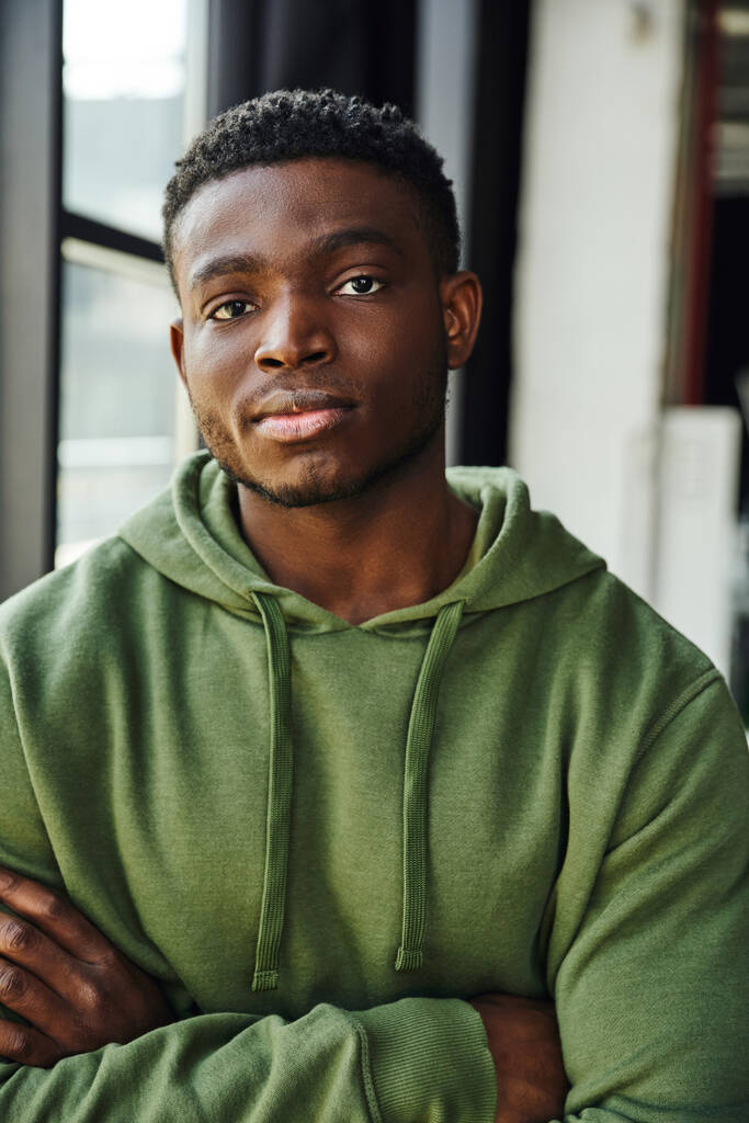 zelfverzekerde en modieuze Afrikaans-Amerikaanse man op zoek naar camera in hedendaagse kantoorruimte, groene trendy hoodie, gevouwen armen, jonge en succesvolle ondernemer - Foto, afbeelding