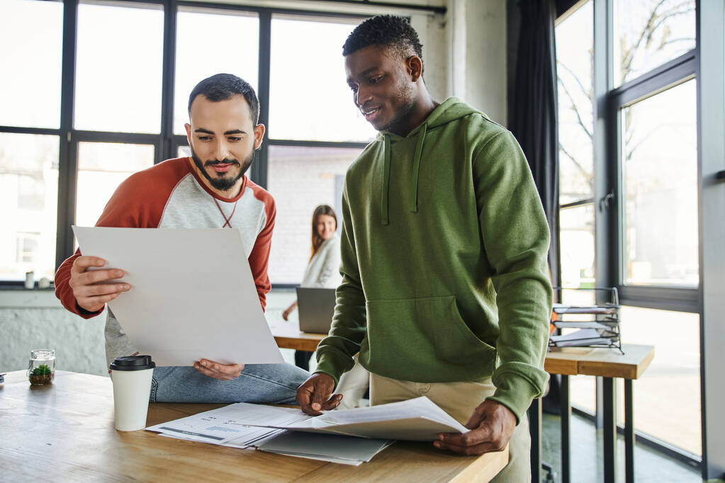 mladí a ambiciózní obchodní partneři pracující na startup plánování v moderní kanceláři, vousatý muž drží dokument v blízkosti afrického amerického kolegy, kreativní myšlení a spolupráci - Fotografie, Obrázek