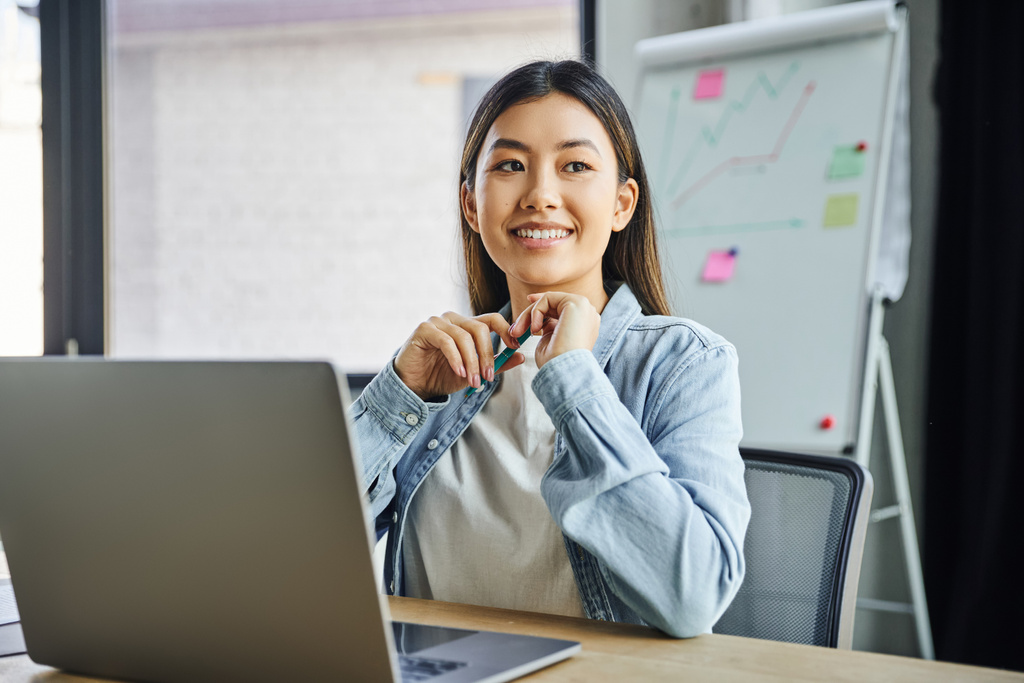 веселая и мечтательная азиатская бизнесвумен в синей джинсовой рубашке сидит перед ноутбуком, держит ручку и смотрит в сторону от флип-чарта на размытом фоне в современном офисе  - Фото, изображение