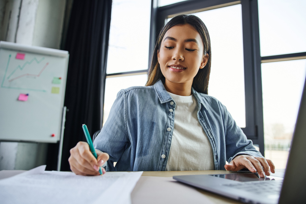 smiling asian woman with brunette hair, in blue denim shirt working on laptop and writing on documents near flip chart on blurred background in contemporary office, successful entrepreneurship - Photo, Image