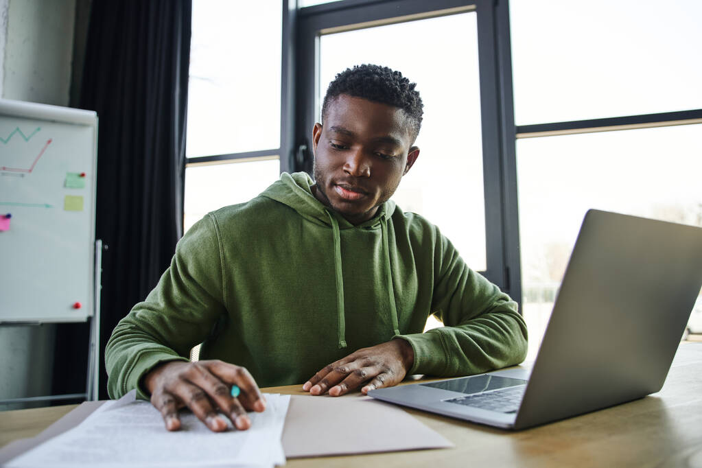 young and stylish african american businessman sitting at workplace and working with documents near laptop and flip chart on blurred background in office, ambitious and career oriented youth - Photo, Image