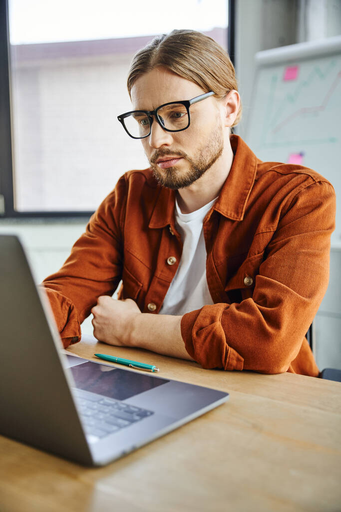 внимательный и концентрированный бородатый предприниматель в модной повседневной одежде и очках, работающий на ноутбуке возле ручки на рабочем столе в современном офисе, деловом образе жизни - Фото, изображение