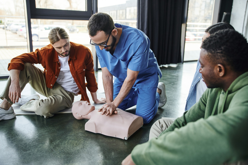 επαγγελματίας νοσηλευτής που κάνει μαλάξεις στο στήθος με CPR manikin και δείχνει καρδιοπνευμονική ανάνηψη σε νέους πολυεθνικούς συμμετέχοντες του σεμιναρίου πρώτων βοηθειών στην αίθουσα εκπαίδευσης - Φωτογραφία, εικόνα