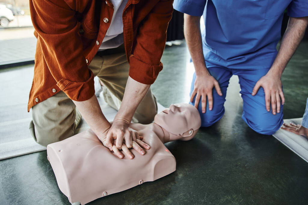 osittainen näkymä nuori mies tekee rintakehän puristuksia CPR-nukke aikana ensiapuseminaarissa lähellä ammatillinen ensihoitaja, kardiopulmonaalinen elvytys, hengenpelastus taitoja ja tekniikoita käsite - Valokuva, kuva