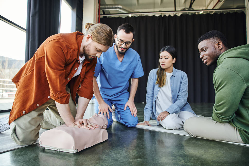 young multiethnic team and professional paramedic looking at african american man doing chest compressions on CPR manikin during first aid hands-on learning, effective life-saving skills concept - Photo, Image