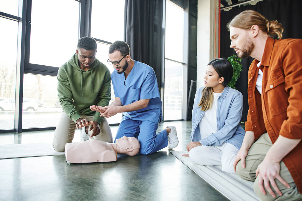 Medizinischer Instruktor erklärt afrikanisch-amerikanischen Mann Herz-Lungen-Wiederbelebungstechniken in der Nähe von CPR-Schaufensterpuppen und jungen multiethnischen Team, lebensrettende Fähigkeiten und Notfallvorsorgekonzept - Foto, Bild