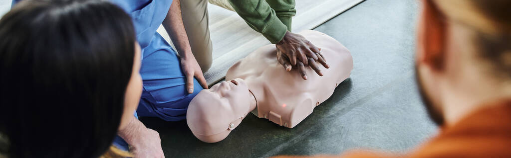 μερική άποψη του Αφροαμερικανού άνδρα που κάνει μαλάξεις στο στήθος στο ανδρείκελο CPR κοντά σε ιατρικό εκπαιδευτή και τους συμμετέχοντες σε θολή πρώτο πλάνο, εξοικονόμηση ζωής δεξιότητες και τεχνικές έννοια, λάβαρο - Φωτογραφία, εικόνα