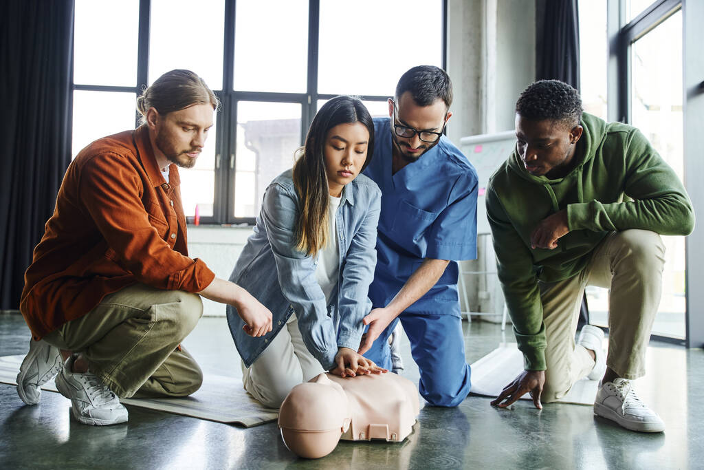 young asian woman doing chest compressions on CPR manikin while practicing cardiopulmonary resuscitation near paramedic and multiethnic participants, life-saving skills and techniques concept - Photo, Image