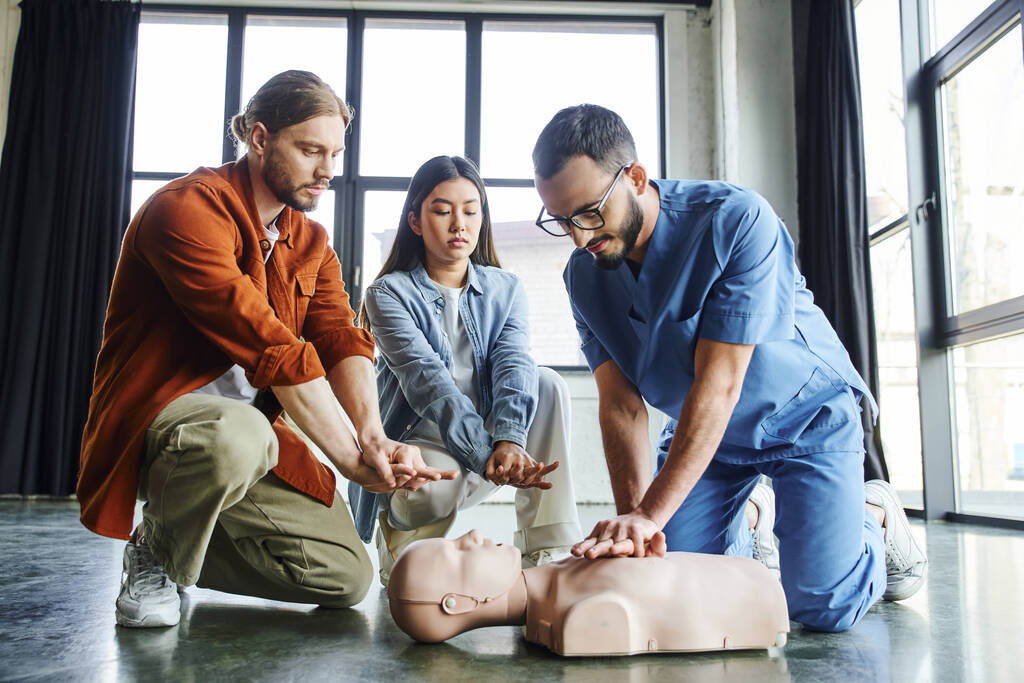 professioneller Rettungssanitäter mit Brille und Uniform, der Brustkompressionen an der CPR-Schaufensterpuppe in der Nähe eines jungen Mannes und einer asiatischen Frau während eines Erste-Hilfe-Trainings zeigt, effektives lebensrettendes Kompetenzkonzept - Foto, Bild