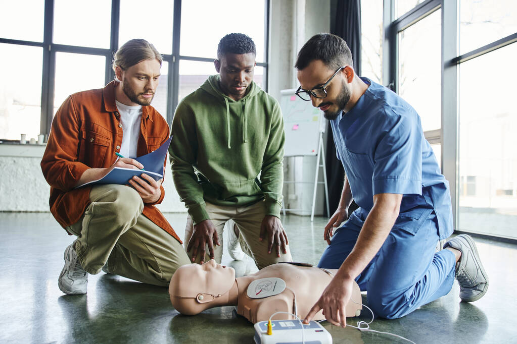 Herz-Wiederbelebung, junger Mann schreibt in Notizbuch in der Nähe eines afrikanisch-amerikanischen Teilnehmers und medizinischer Instruktor bedient Defibrillator an CPR-Schaufensterpuppe während eines Erste-Hilfe-Trainings - Foto, Bild