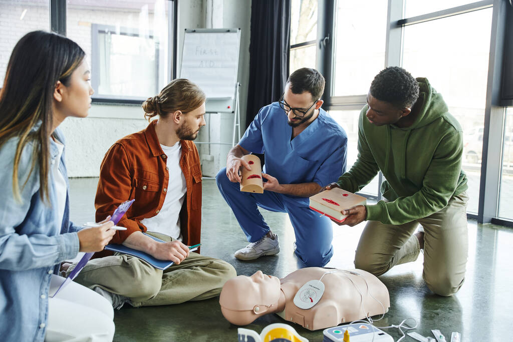 medical instructor and african american man holding wound care simulators near CPR manikin, defibrillator and multiethnic participants with notebook and clipboard, life-saving skills concept - Photo, Image