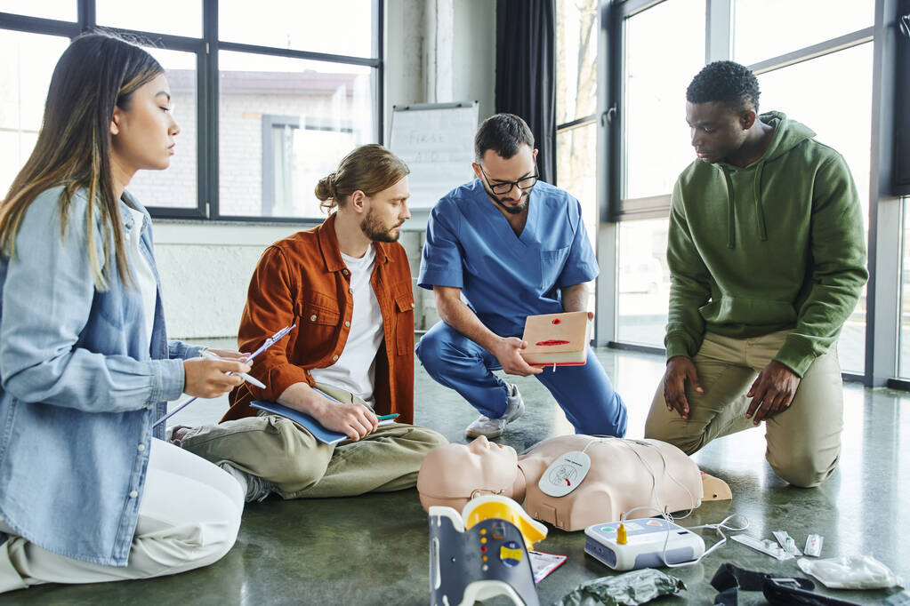 CPR manikin, defibrilatör ve tıbbi ekipman yakınlarındaki çok kültürlü ekipmanlara ilk yardım ve hayat kurtarma konsepti sunan uzman sağlık görevlileri - Fotoğraf, Görsel