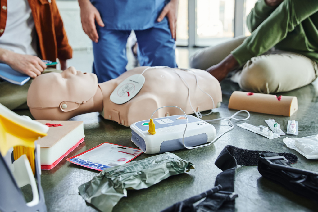 CPR manikin, дефібрилятор, симулятори догляду за ранами, компресійний турнікет, бинти та шприци поблизу інструктора та багатоетнічних учасників семінару першої допомоги, концепція екстреної готовності
 - Фото, зображення