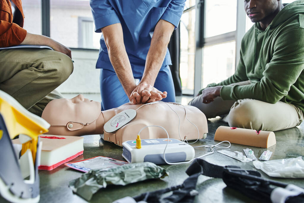 μερική άποψη του νοσηλευτή που κάνει μαλάξεις στο στήθος για CPR manikin κοντά σε πολυεθνικούς συμμετέχοντες και ιατρικό εξοπλισμό, απινιδωτή, προσομοιωτές περιποίησης τραυμάτων, αιμοστατικοί επίδεσμοι, σεμινάριο εκπαίδευσης πρώτων βοηθειών - Φωτογραφία, εικόνα