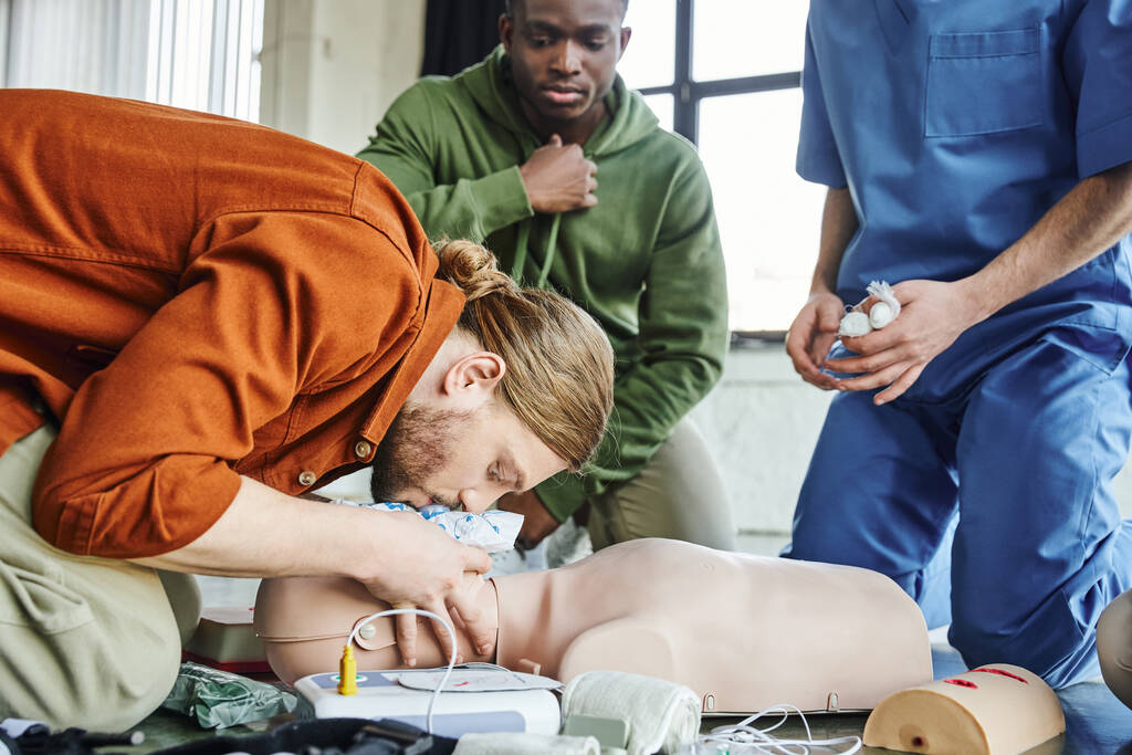 EHBO-seminar, jonge man die levensreddende vaardigheden beoefent en kunstmatige ademhaling doet aan reanimatiepop bij defibrillator, medische apparatuur en Afrikaans-Amerikaanse man met medische instructeur - Foto, afbeelding