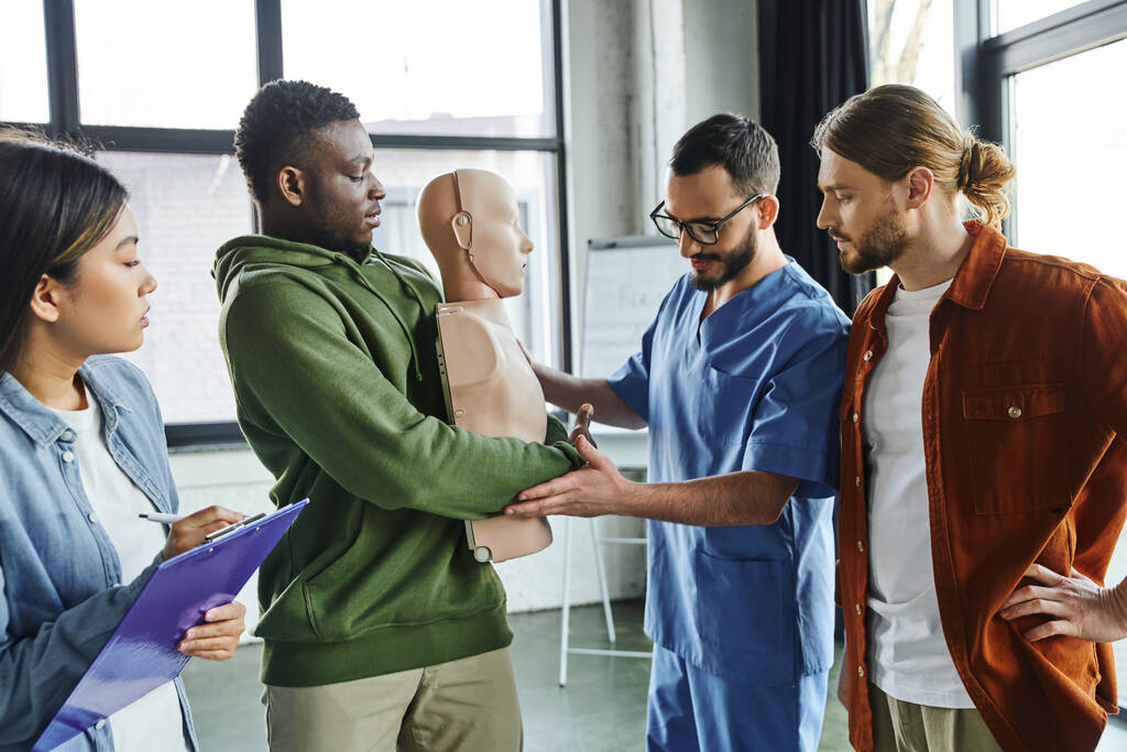medical instructor assisting african american man practicing life-saving techniques in case of choking on CPR manikin near multiethnic participants, emergency situations preparedness concept - Photo, Image