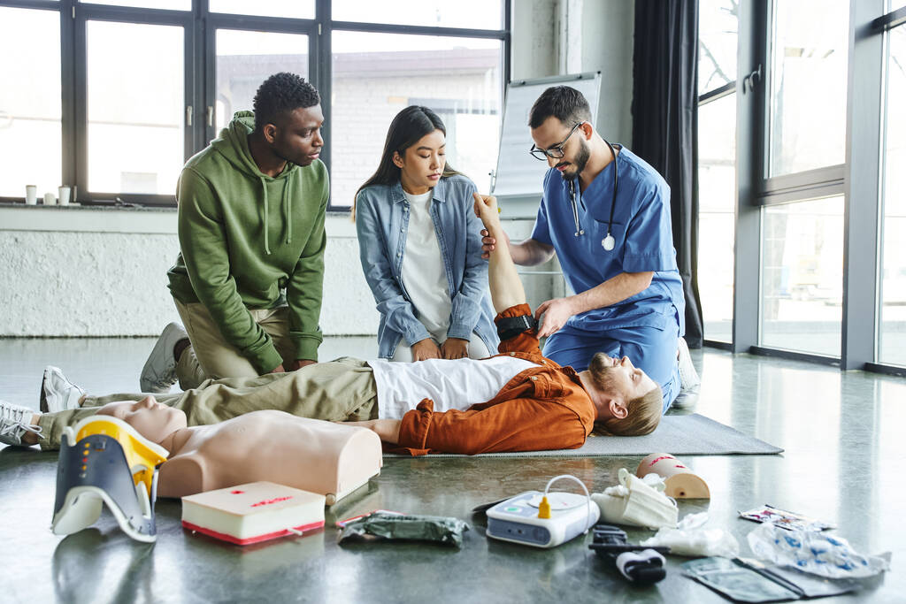 medical instructor applying compression tourniquet on arm of seminar participant near asian woman, african american man and medical equipment in training room, emergency situations response concept - Photo, Image