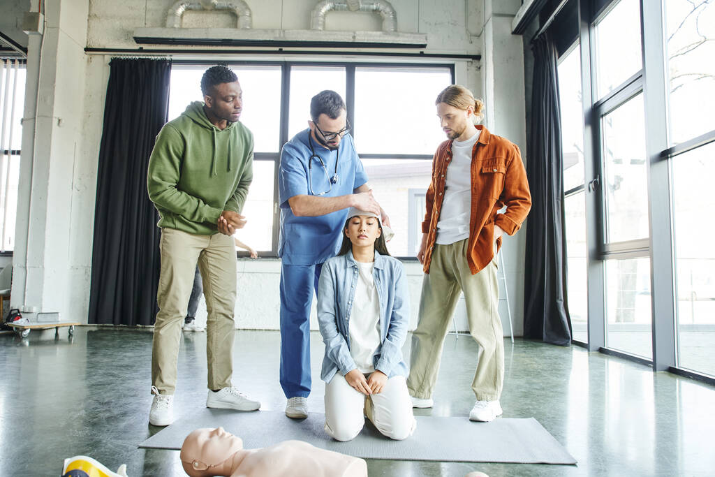 πολυεθνικός άνδρας που εξετάζει επαγγελματία τραυματιοφορέα επιδέσμους επικεφαλής της ασιατικής γυναίκας κοντά στο ανδρείκελο CPR κατά τη διάρκεια σεμιναρίου κατάρτισης πρώτων βοηθειών, σημασία της έννοιας της ετοιμότητας έκτακτης ανάγκης - Φωτογραφία, εικόνα