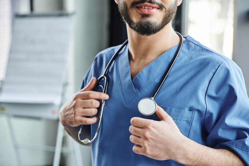 άποψη του θετικού γενειοφόρου γιατρού με μπλε στολή που αγγίζει στηθοσκόπιο στο λαιμό, ενώ στέκεται στο δωμάτιο κατάρτισης, σεμινάριο κατάρτισης πρώτων βοηθειών και έννοια ετοιμότητας έκτακτης ανάγκης - Φωτογραφία, εικόνα