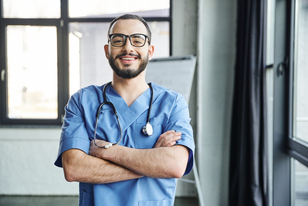 fröhlicher, bärtiger Arzt mit Brille und blauer Uniform, der mit verschränkten Armen und Stethoskop vor Kamera, Erste-Hilfe-Seminar und Notfallpräventionskonzept steht - Foto, Bild