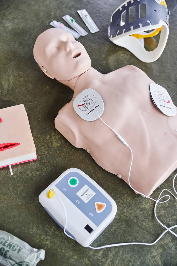 high angle view of CPR manikin, external defibrillator, wound care simulator, neck brace and syringes on floor in training room, medical equipment for first aid training and skills development - Photo, Image