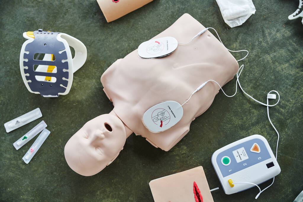Draufsicht auf die CPR-Schaufensterpuppe in der Nähe automatisierter Defibrillatoren, Wundbehandlungssimulatoren, Nackenstütze und Spritzen auf dem Boden im Schulungsraum, medizinische Ausrüstung für Erste-Hilfe-Schulungen und Kompetenzentwicklung - Foto, Bild