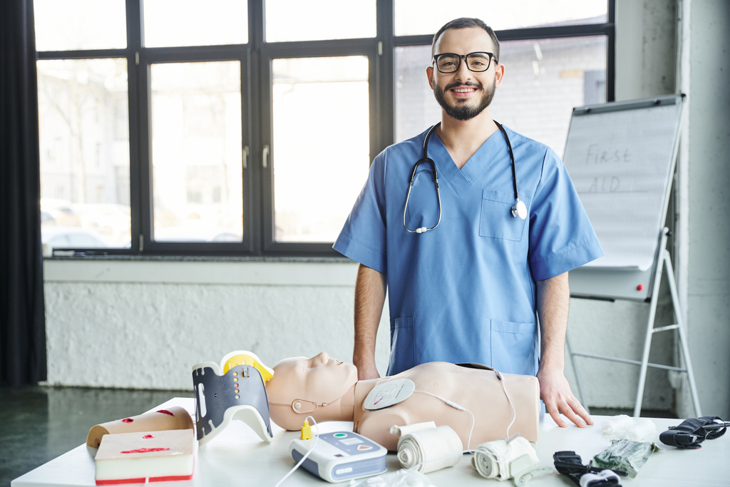 joyful medical instructor looking at camera near CPR manikin, defibrillator, compressive bandages and tourniquets in training room, first aid hands-on learning and critical skills development concept - Photo, Image