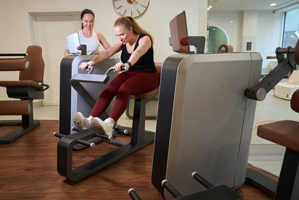 Cliente de clínica trabalha em uma máquina de fitness moderna, ao lado dele é um treinador-consultor - Foto, Imagem