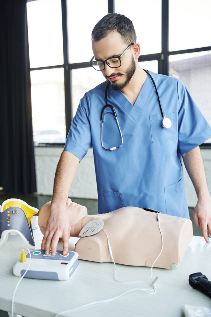 healthcare worker in blue uniform, stethoscope and eyeglasses operating automated defibrillator near CPR manikin, first aid hands-on learning and critical skills development concept - Photo, Image