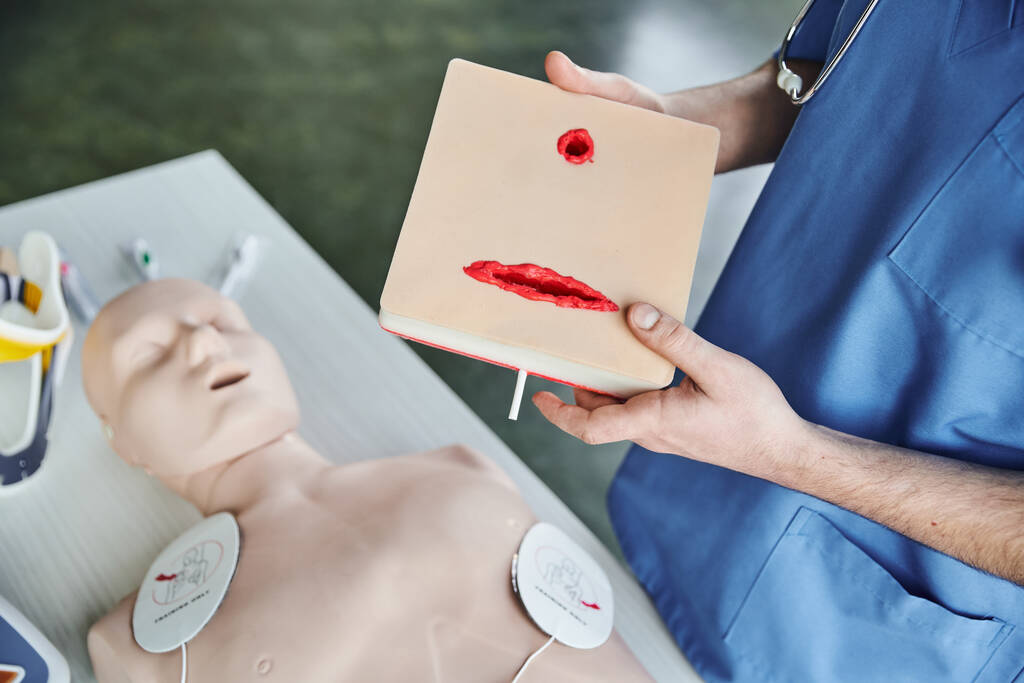 partial view of medical instructor in blue uniform holding wound care simulator above CPR manikin with defibrillator pads, first aid hands-on learning and critical skills development concept - Photo, Image