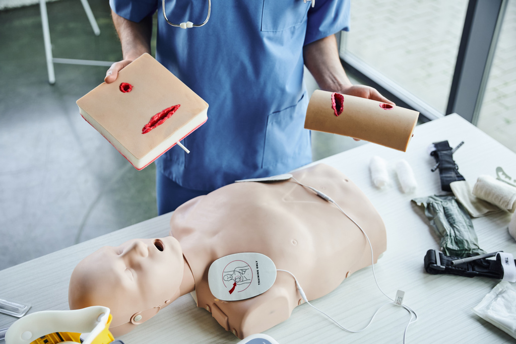 częściowy widok instruktora medycznego stojącego z symulatorem pielęgnacji ran w pobliżu manekina resuscytacyjnego z defibrylatorem i sprzętem medycznym, praktyczna nauka pierwszej pomocy i koncepcja rozwoju umiejętności krytycznych - Zdjęcie, obraz