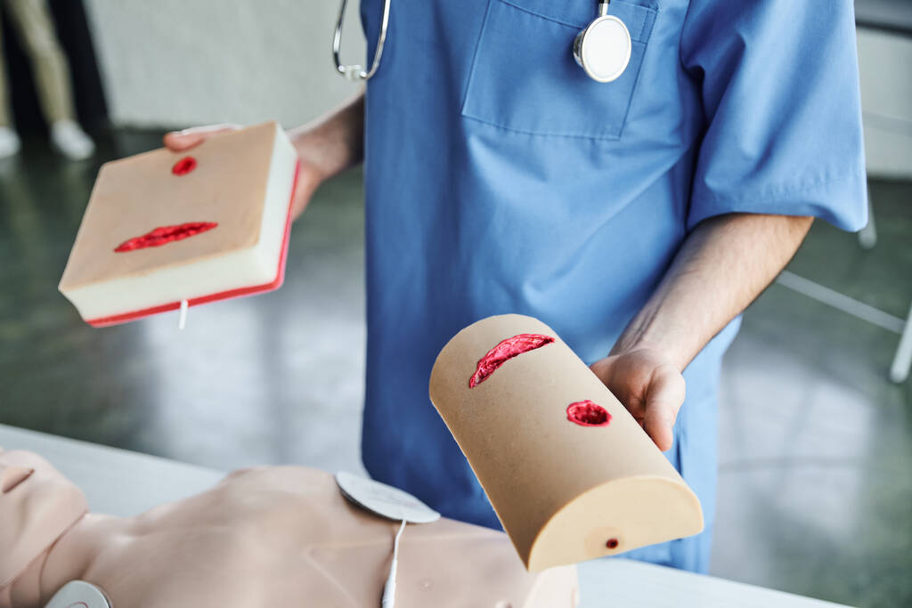 częściowy widok instruktora medycznego pokazującego symulatory pielęgnacji ran stojącego w pobliżu manekina resuscytacyjnego w sali szkoleniowej, praktyczną naukę pierwszej pomocy oraz koncepcję rozwoju umiejętności krytycznych - Zdjęcie, obraz