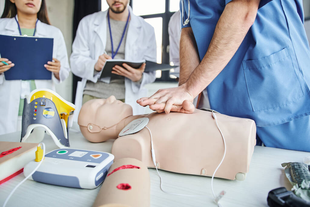 partial view of paramedic practicing chest compressions on CPR manikin near defibrillator, neck brace and students with clipboard and notebook, life-saving skills hands-on learning concept - Photo, Image
