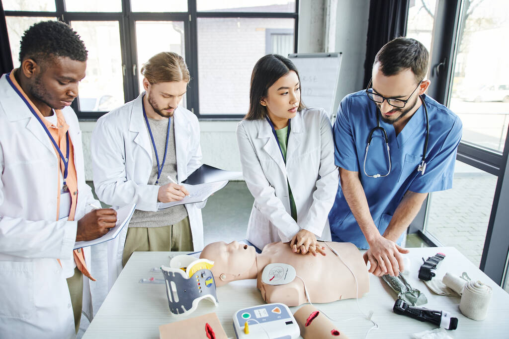 multiethnic students in white coats writing in notebooks near medical instructor assisting asian woman doing chest compressions on CPR manikin, emergency situations response concept - Photo, Image